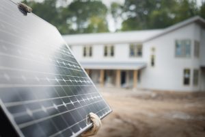 	
A person wearing work gloves holds a solar panel in front of a new house under construction on a dirt lot, highlighting Solar Panel Installation Lafourche Parish.