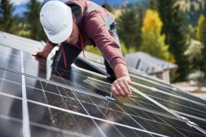 A worker in a hard hat, representing Solar Installers in St Charles Parish, adjusts a solar panel outdoors using a tool, with trees and rooftops visible in the background.