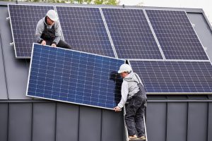 Two workers wearing helmets install solar panels on the sloped roof of a building, with one worker on a ladder and the other on the roof—showcasing skilled Solar Installers in St Charles Parish at work.